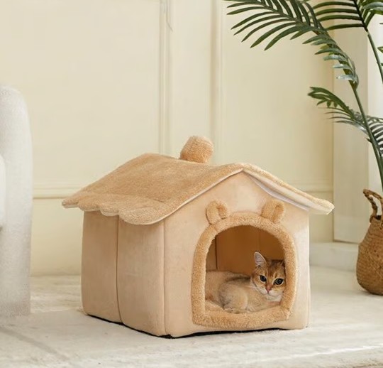 Kat ligt ontspannen in beige kattenhuisje met oortjes en golvend dak, in een rustige woonkamer.